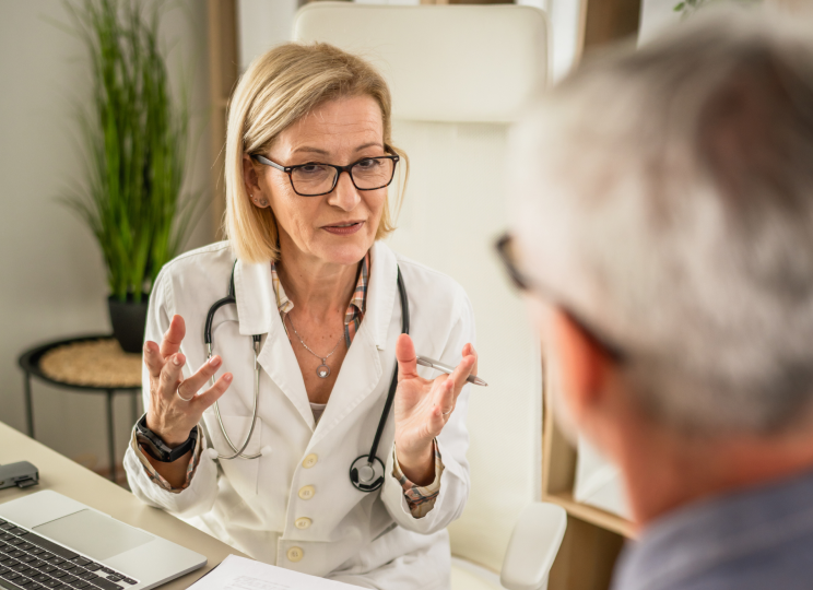 A medical professional in a white coat sitting at a desk, talking with another person across from her.