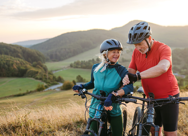 Two people wearing helmets, standing outdoors with bicycles on a hillside.