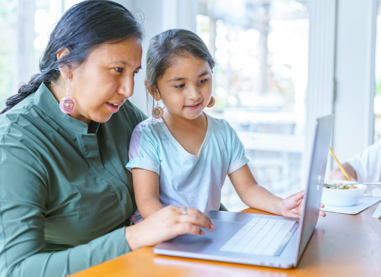 An adult and a child sitting together indoors, looking at a laptop screen.
