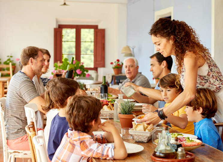 A group of adults and children gathered around a dining table indoors, with one woman standing and serving food.