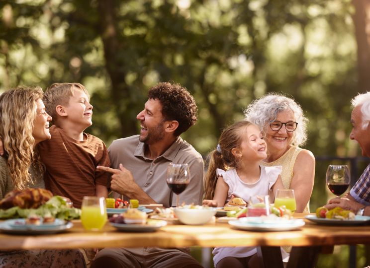 A group of adults and children sitting together at an outdoor table, talking and eating.